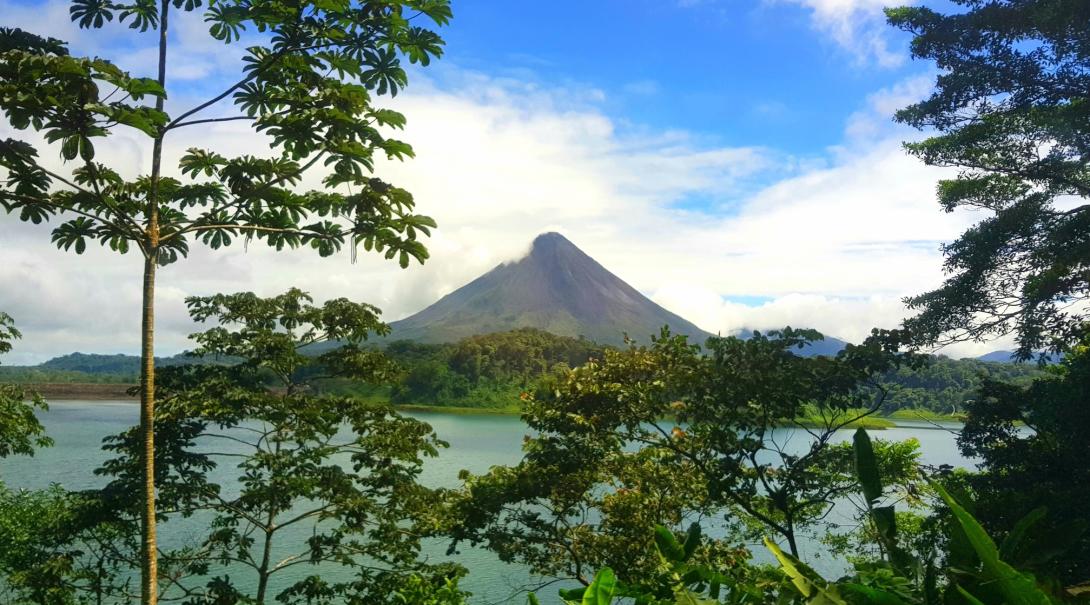 Arenal Volcano standing tall in the distance on a sunny day A stunning view of Arenal Volcano shot through the overgrowth in Costa Rica, showing bright green fauna and a pyramid peak volcano